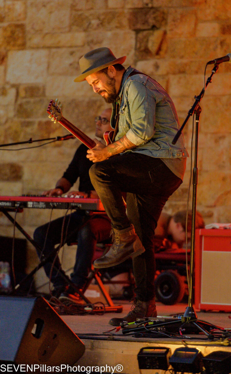 Man in a light brown fedora lifting his leg up while playing guitar on stage