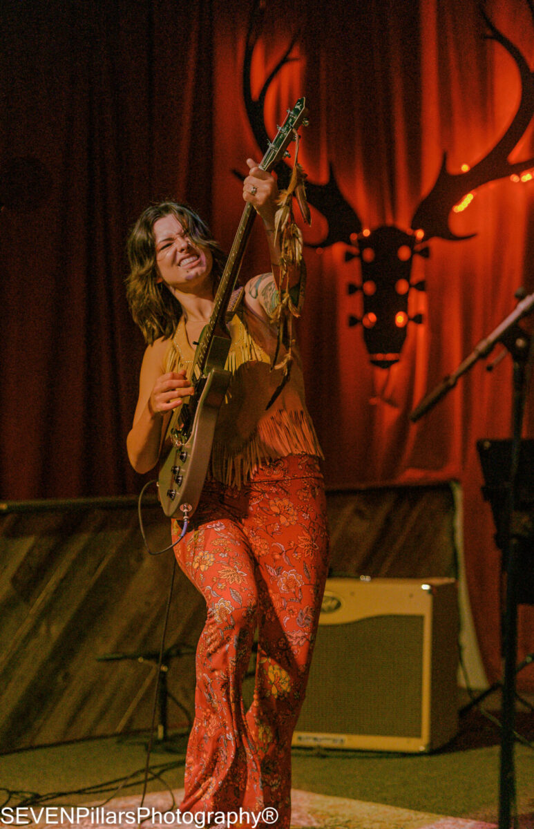 Woman in a yellow-and-orange outfit playing a guitar in front of a orange curtain with a moose silhouette decor