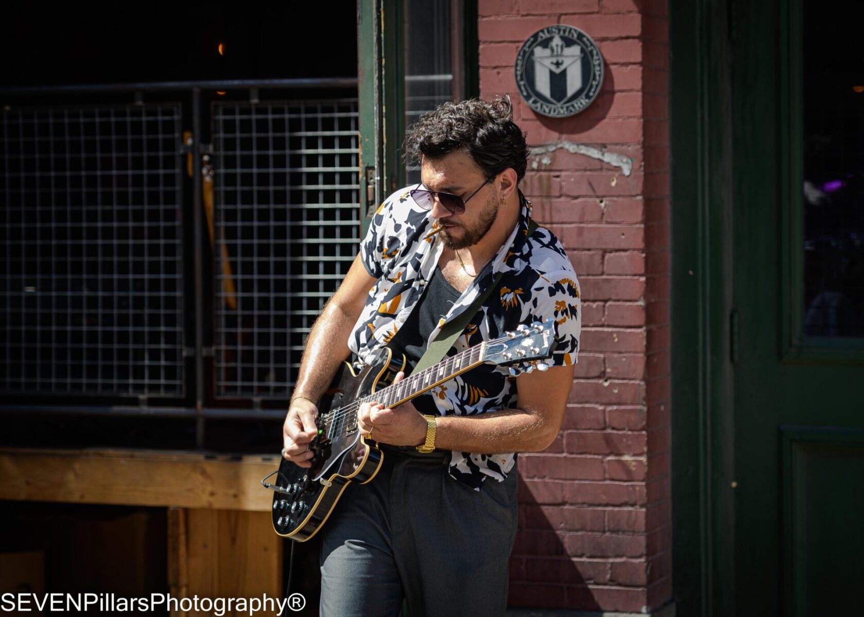 Man wearing sunglasses playing a black electric guitar in front of an establishment